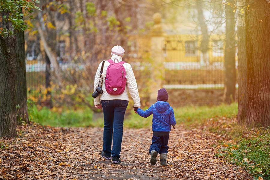 A woman and a small child hold hands while walking through a park, seen from behind