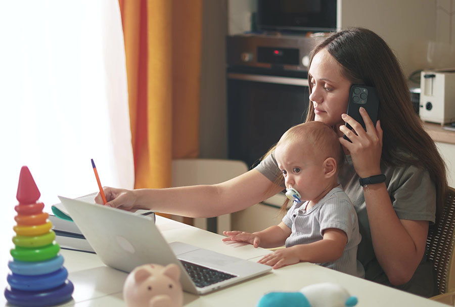 A mother sits at a dining table with a baby on her lap, holding a phone and with an open laptop in front of her