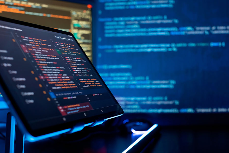 laptop screen filled will lines of code, with a large screen filled with code next to it