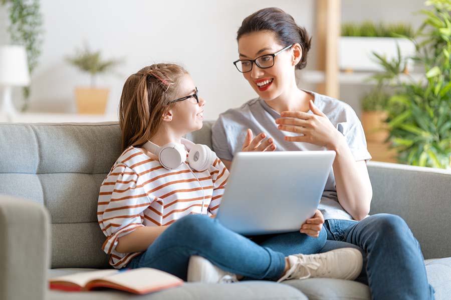 A smiling mother and young daughter have a conversation on a sofa, using a laptop