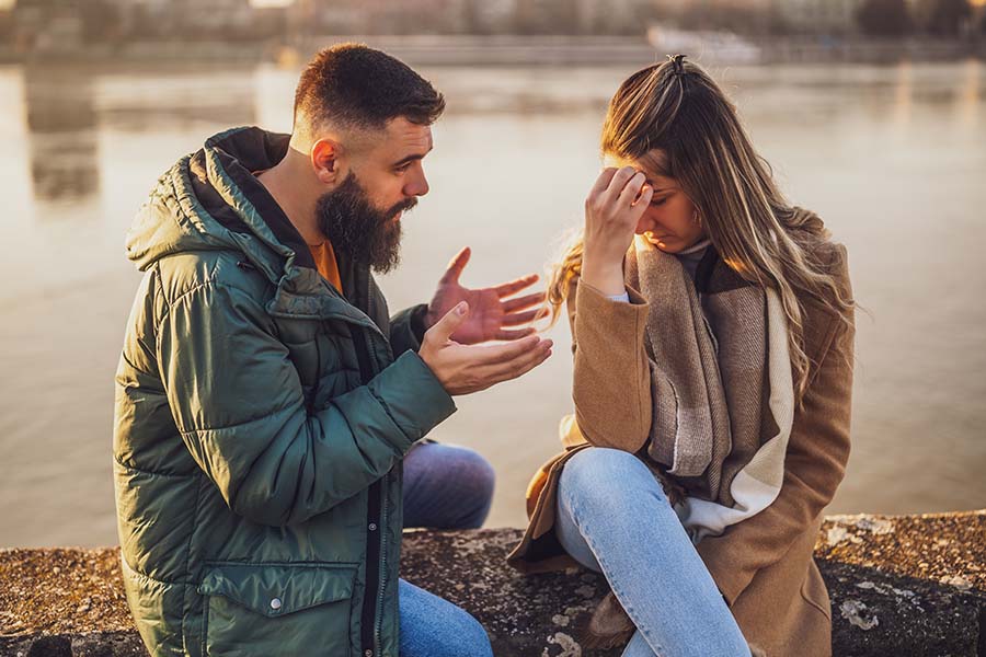 A couple in warm clothes sit by a lake appearing to be deep in an argument
