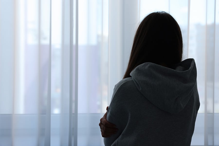 A woman viewed from behind stands in front of a window covered by curtains