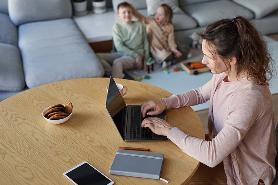 A tired looking woman uses a laptop as two children fight in the background