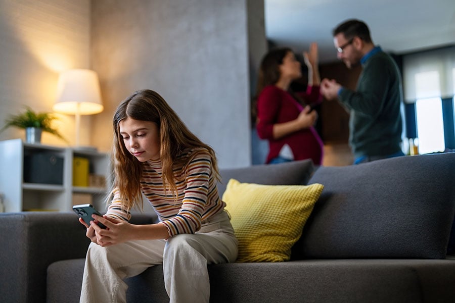 A teen sits on a sofa using a phone while adults argue in the background