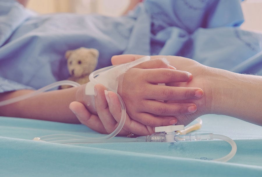 A close-up of an adult holding a child's hand in a hospital bed. The child has a cannula in their hand.