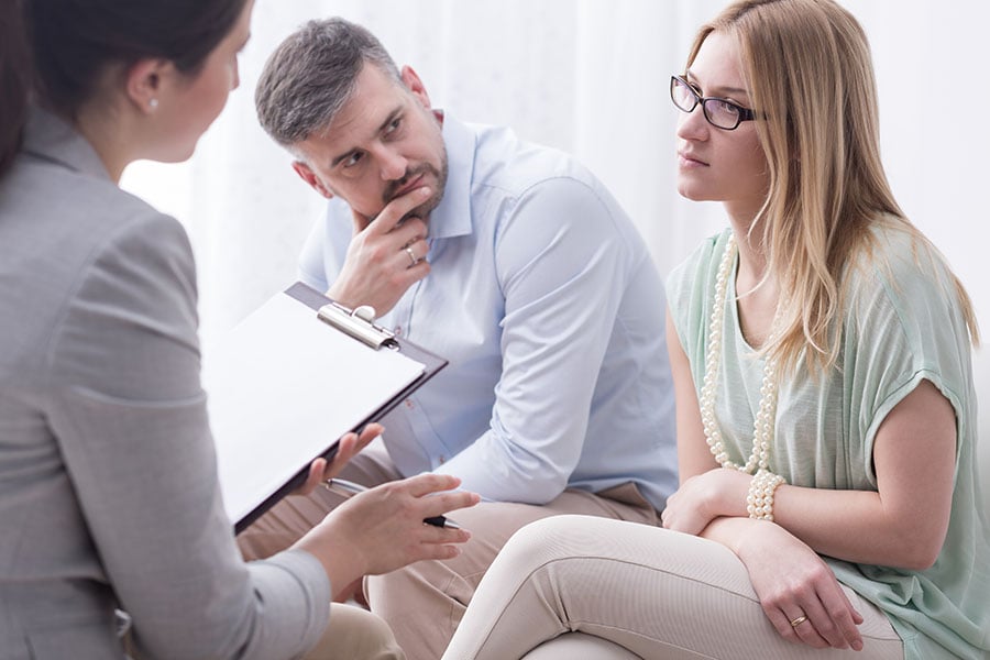 A young couple sit together on a couch in discussion with a professional woman holding a clipboard