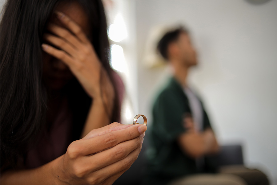 A woman in the foreground holding a ring, with a man in the background with arms folded