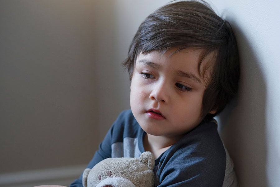 A young boy around toddler age sits alone cuddling a teddy looking sad