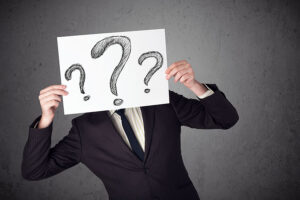 A man in a black suit holds up a piece of paper with large questions marks drawn on it