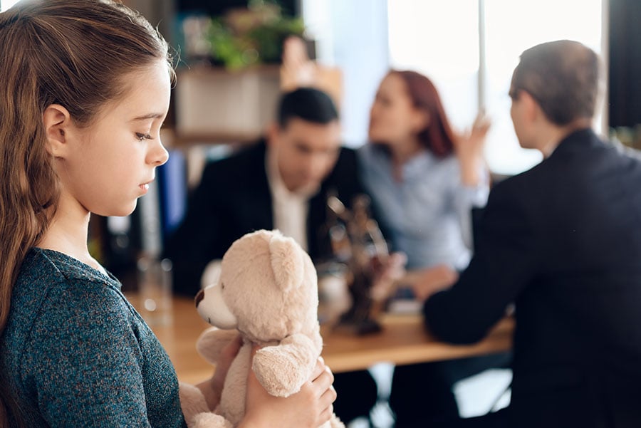 A young girl holding a teddy in the foreground with a couple arguing in front of a judge or solicitor in the background