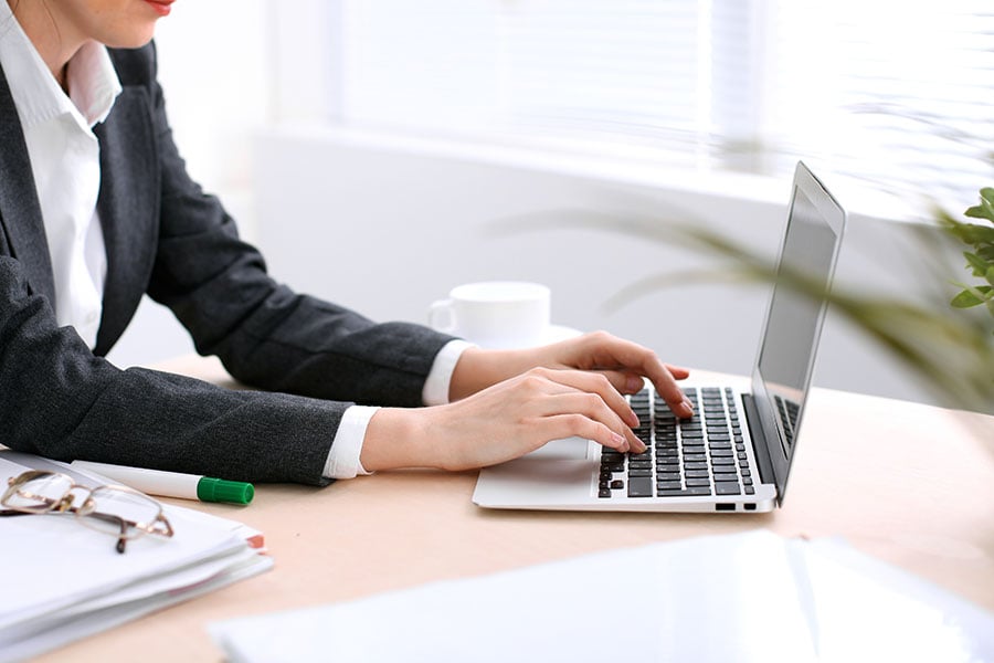 A woman in a black suite working on a laptop with a notepad and pen next to her