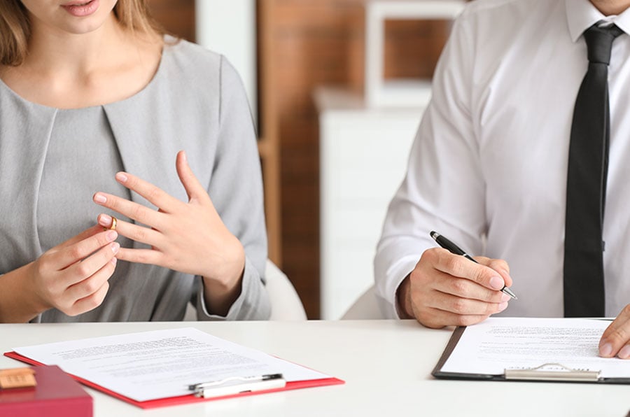A woman taking off a wedding ring sits next to a man signing a form